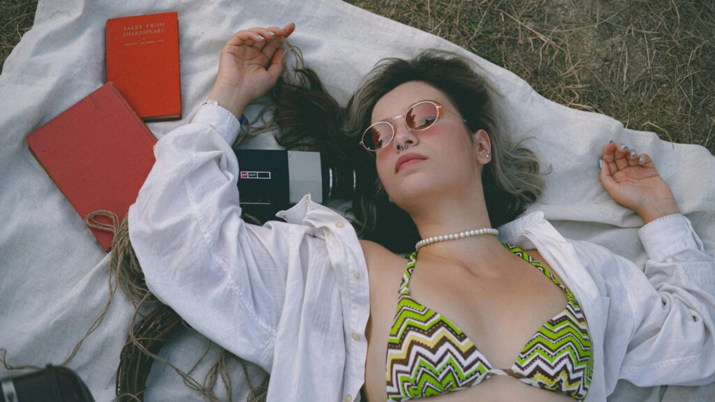 Woman in a bikini and sunglasses relaxing on a blanket outdoors with books and vintage camera.